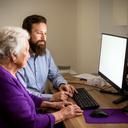 Eanna Mörsch, patient tech tutor with beard, sitting beside an elderly student at a desktop computer